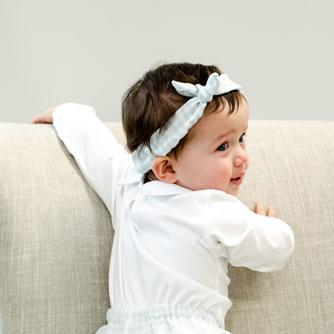 A newborn with dark hair wearing the Isla Tie Headband and white outfit looks over her shoulder, smiling, while lying on a beige sofa.