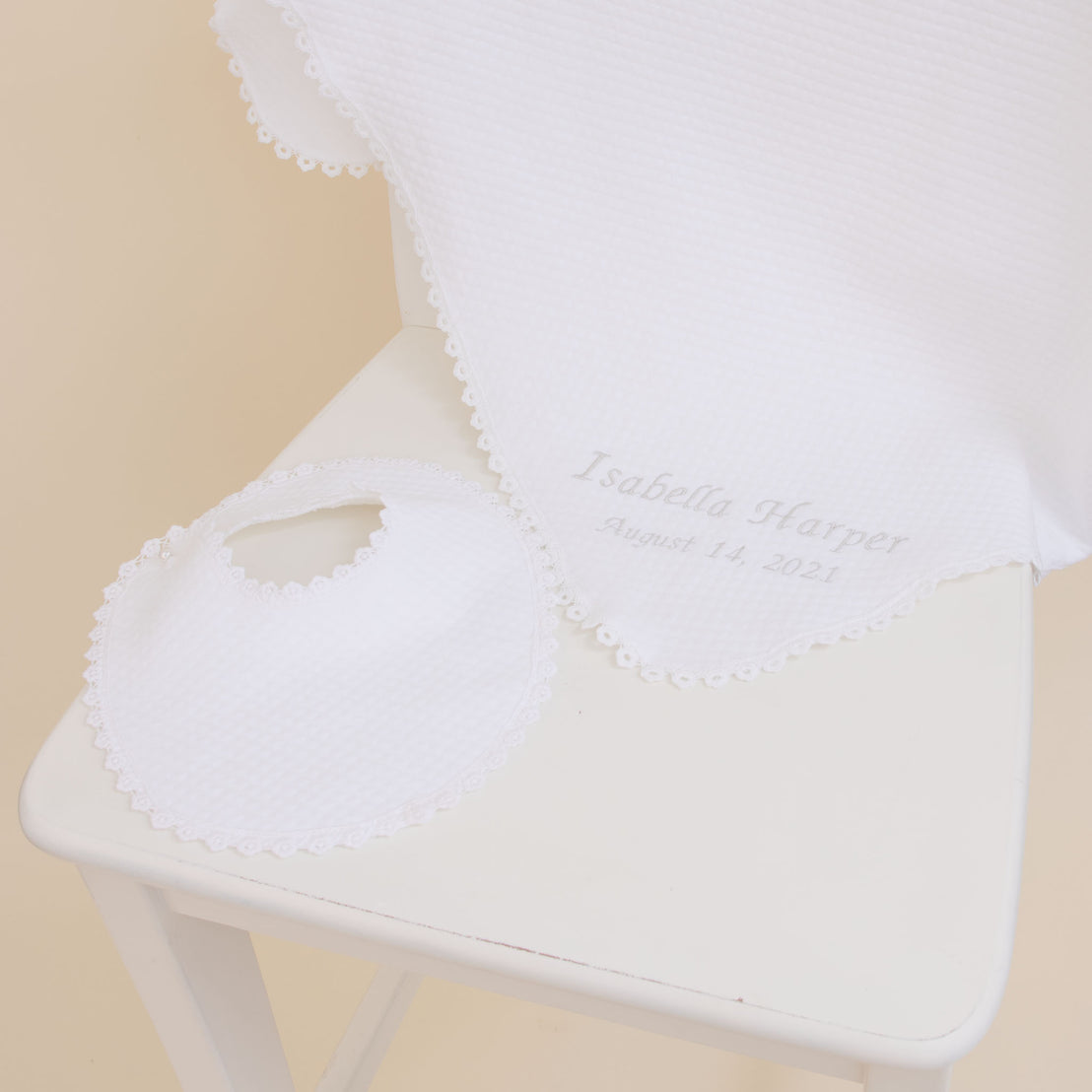 A Baby Beau & Belle Baby Girl Personalized Gift Set, consisting of a white quilted cotton bib and blanket embroidered with the name "Isabella Harper August 14, 2021," displayed on a white chair against a beige background.
