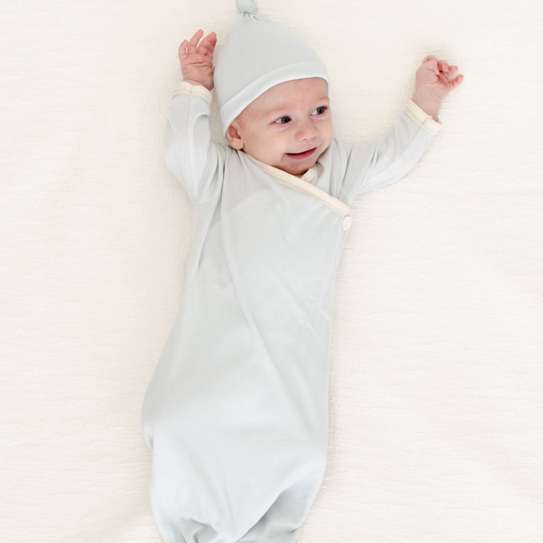 A newborn boy, dressed in the Evan Knot Gown with a matching hat, lies joyfully on a soft surface. This delightful outfit is expertly handmade in the USA from 100% pima cotton, ensuring both comfort and style as he stretches his arms above his head with a smile.