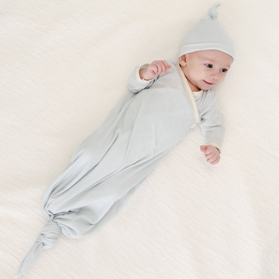 A baby lies on a soft white blanket, smiling and looking upward, dressed in the Evan Knot Gown—a light gray newborn boys outfit handmade in the USA from 100% pima cotton—with a matching hat.