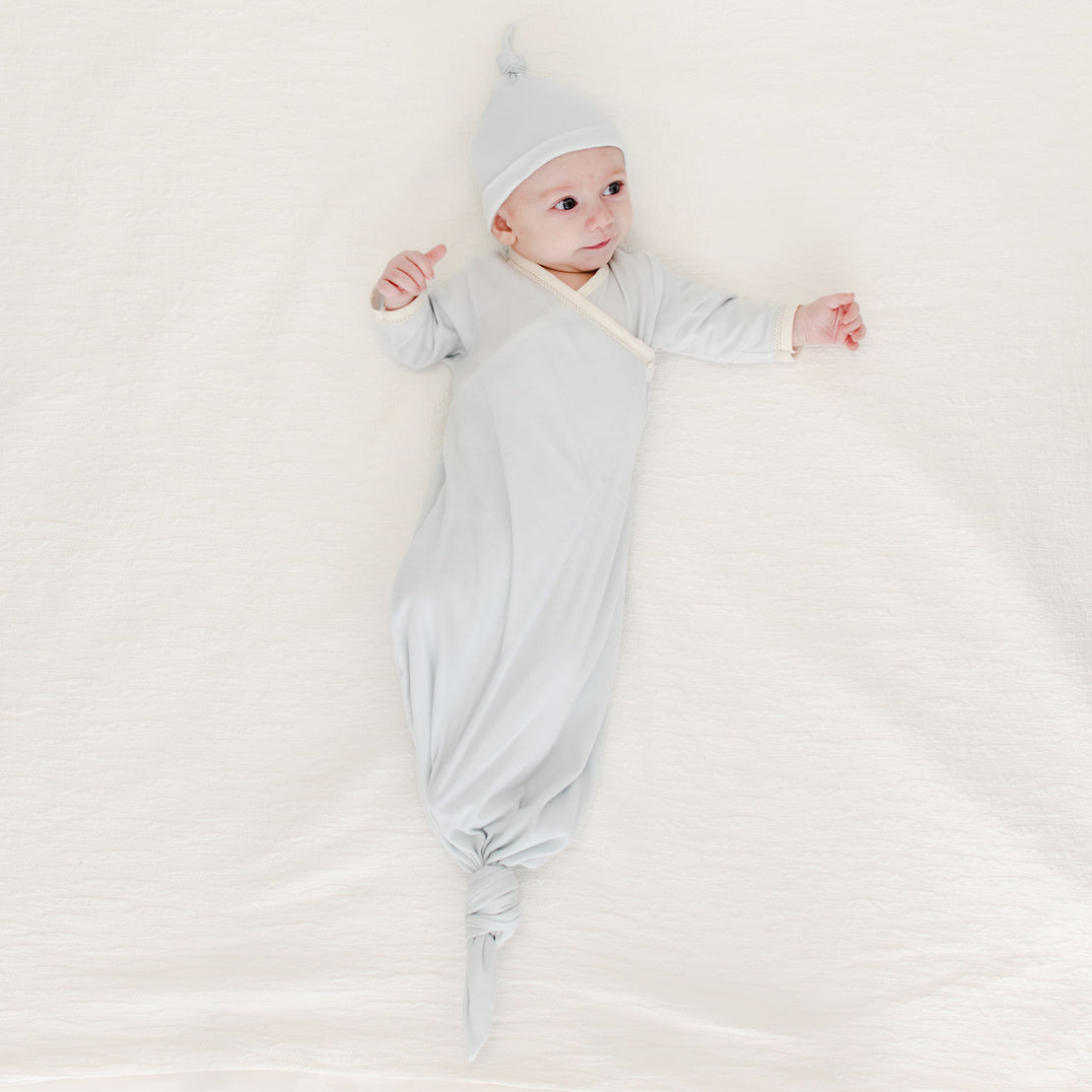 A baby in the Evan Knot Gown, artisanally crafted from 100% pima cotton, wears a coordinating hat while reclining on a cozy, cream-colored surface. The baby looks upward with arms gently outstretched. Skillfully made in the USA with love and care.