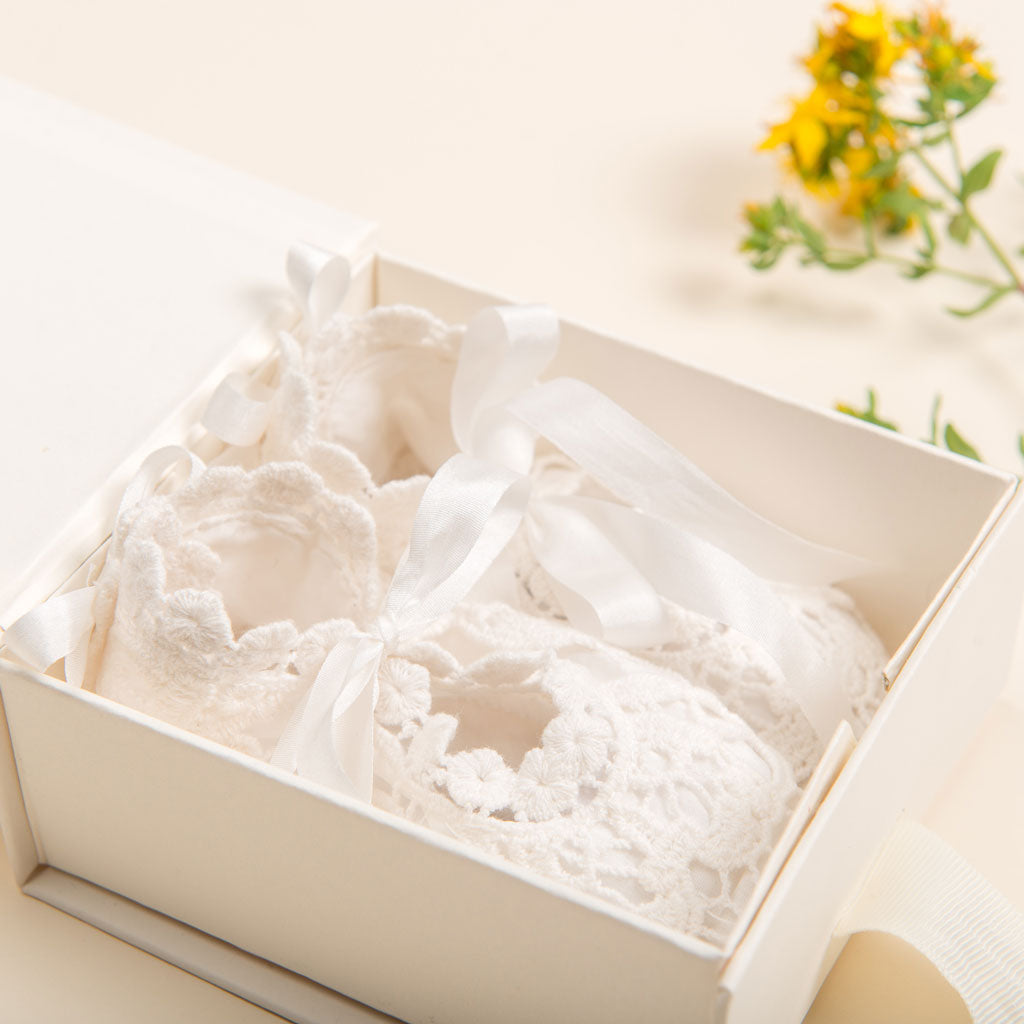 A pair of delicate white baby booties featuring lace and ribbon embellishments is elegantly presented in an open cream-colored mini gift box, adorned with a soft ivory tissue lining. A small yellow flower in the background adds a touch of color, enhancing this charming scene by Baby Beau & Belle.
