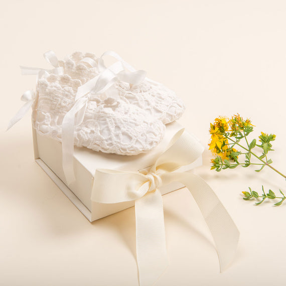 White crocheted baby booties from Baby Beau & Belle, decorated with satin ribbons, are gracefully placed on a cream gift box known as "Gift Wrap | Mini Gift Box with Bow," elegantly encased in ivory tissue. A small branch adorned with yellow flowers completes the charming display against a light cream background.