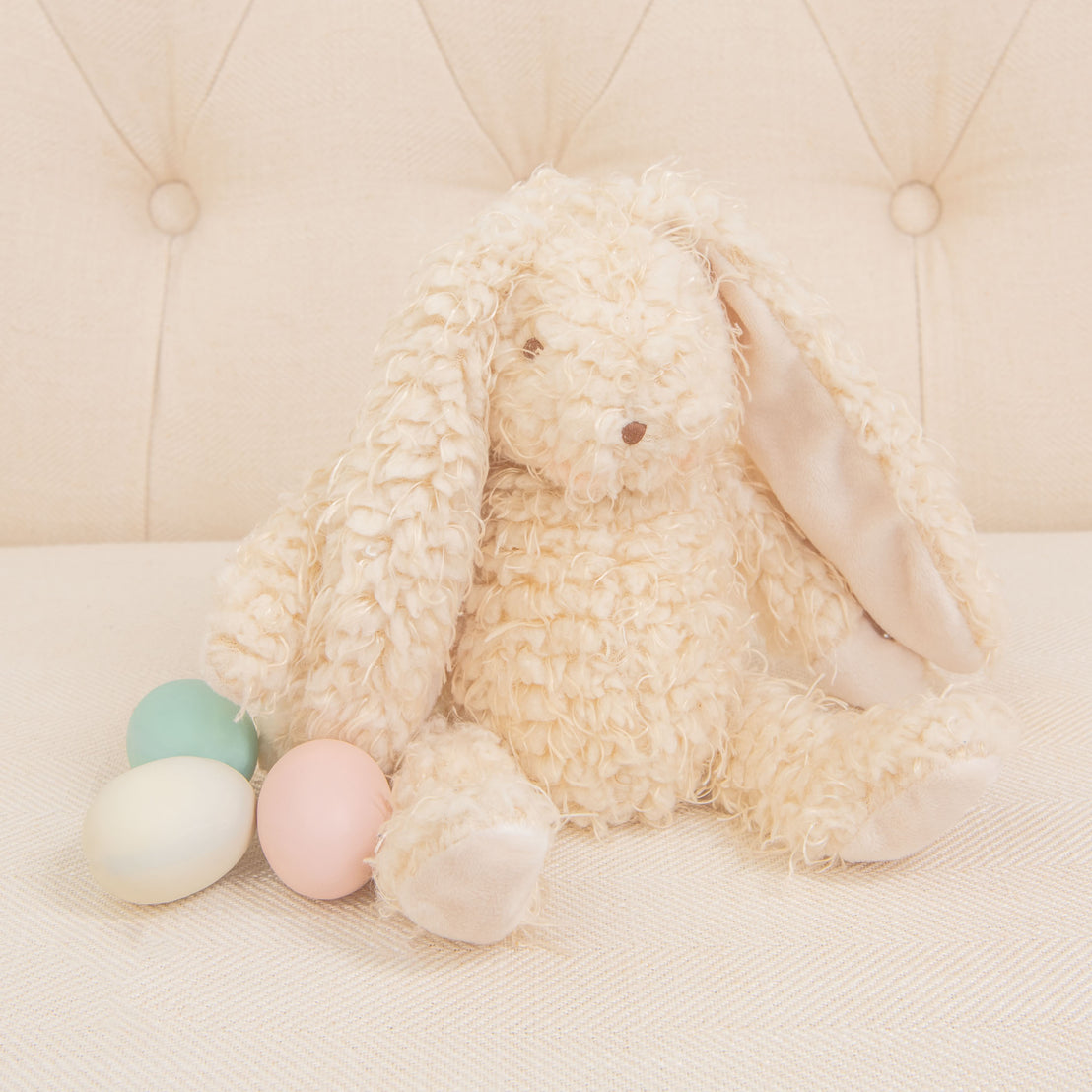 A plush Harey Bunny, featuring cream-colored and soft almond scraggle fur, is sitting on a light-colored sofa. With its long ears, it's joined by three pastel-colored eggs—green, pink, and off-white. This delightful arrangement serves as an ideal baby gift against the tufted upholstery backdrop.