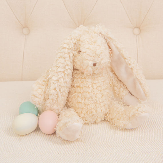 A plush Harey Bunny, featuring cream-colored and soft almond scraggle fur, is sitting on a light-colored sofa. With its long ears, it's joined by three pastel-colored eggs—green, pink, and off-white. This delightful arrangement serves as an ideal baby gift against the tufted upholstery backdrop.