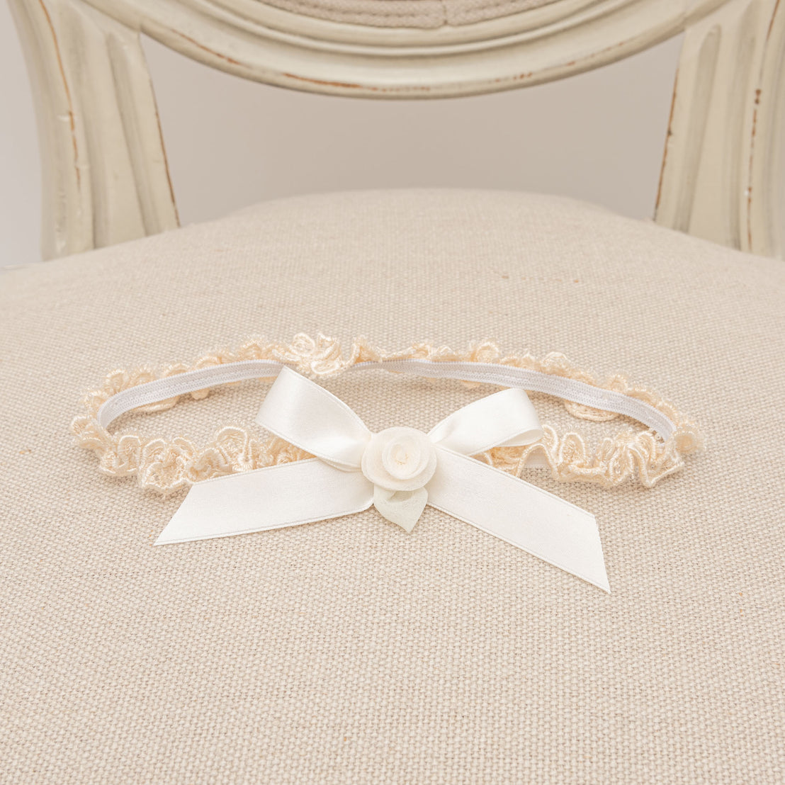 An elegant Kristina Headband with a white satin bow and a delicate white rose, presented on a cushioned chair with creamy upholstery and intricate frame design. Perfect for a baptism or christening.