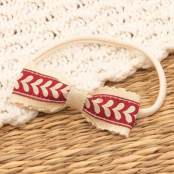 The Valentina Headband, featuring a handmade beige band with a red and white patterned bow, rests on a woven surface. It's an ideal accessory for adding charm to any holiday outfit and comes with textured white fabric.