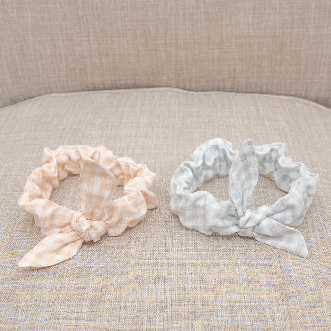 Two Isla Tie Headbands on a beige sofa, one plain peach and the other light blue checkered, both with decorative knots in a vintage style.