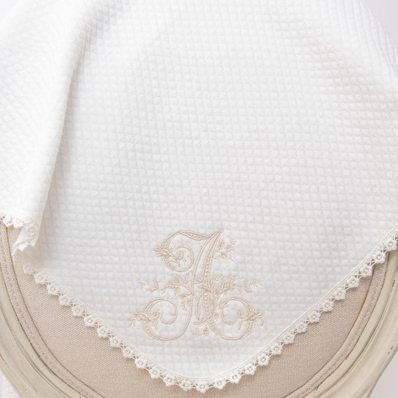Elegant white baby blankets with initial embroidery personalization 'a' and delicate lace trim, draped over the edge of a soft beige cushioned chair.