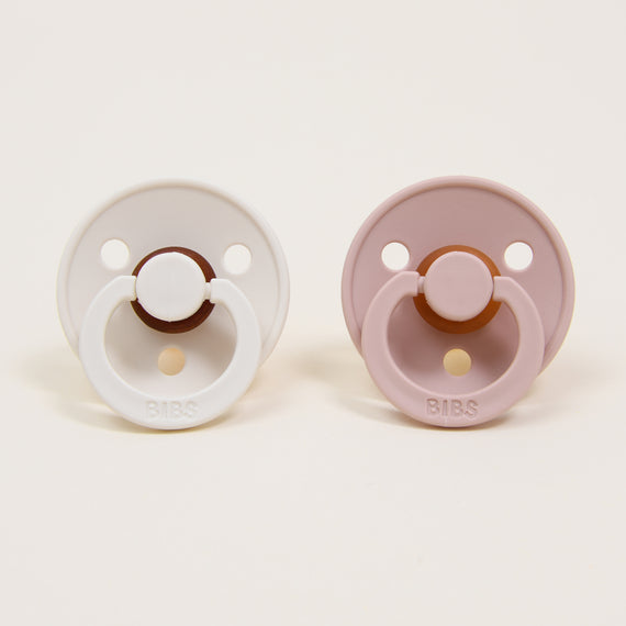 Two Bibs Pacifiers placed on a white background, one white and one mauve, both with round handles and three ventilation holes.