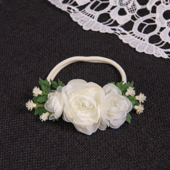 The Guinevere Flower Headband, adorned with ivory flowers and green leaves, rests on a dark fabric surface. Nearby, a piece of white lace subtly enhances the delicate ambiance.