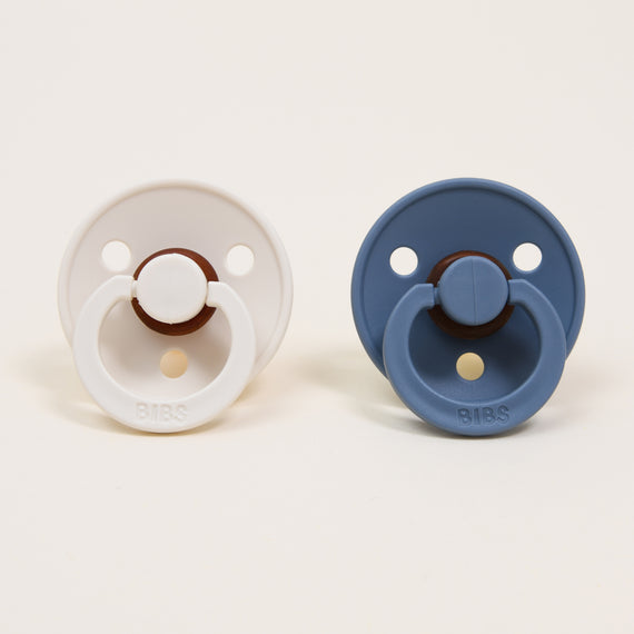 Two Bibs Pacifiers side-by-side on a plain background; one is white and the other is blue, set against a white background.