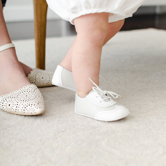 Baby boy wearing the Ivory Two Tone Wingtip Shoes that match the Oliver Baby Romper. They are made with a white patent leather and ivory matte leather with detailed edging. But bottoms are made with ivory suede leather.