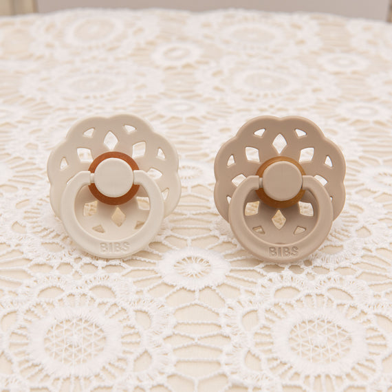 Pair of scalloped pacifiers in vanilla and ivory tones, displayed on a floral lace background, showcasing their delicate design.