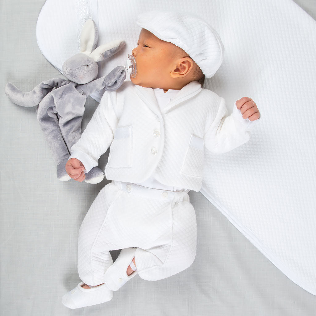 A baby wearing the Elijah Newborn 3-Piece Suit and matching cap lies on a soft, white surface next to a gray stuffed rabbit, serenely holding a pacifier in their mouth.