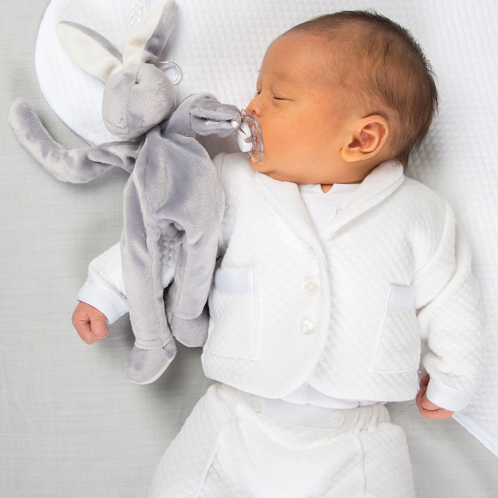 A sleeping baby, dressed in the Elijah Newborn 3-Piece Suit, cuddles a gray plush bunny while holding a pacifier in its mouth. The little one is lying on a soft, white surface.