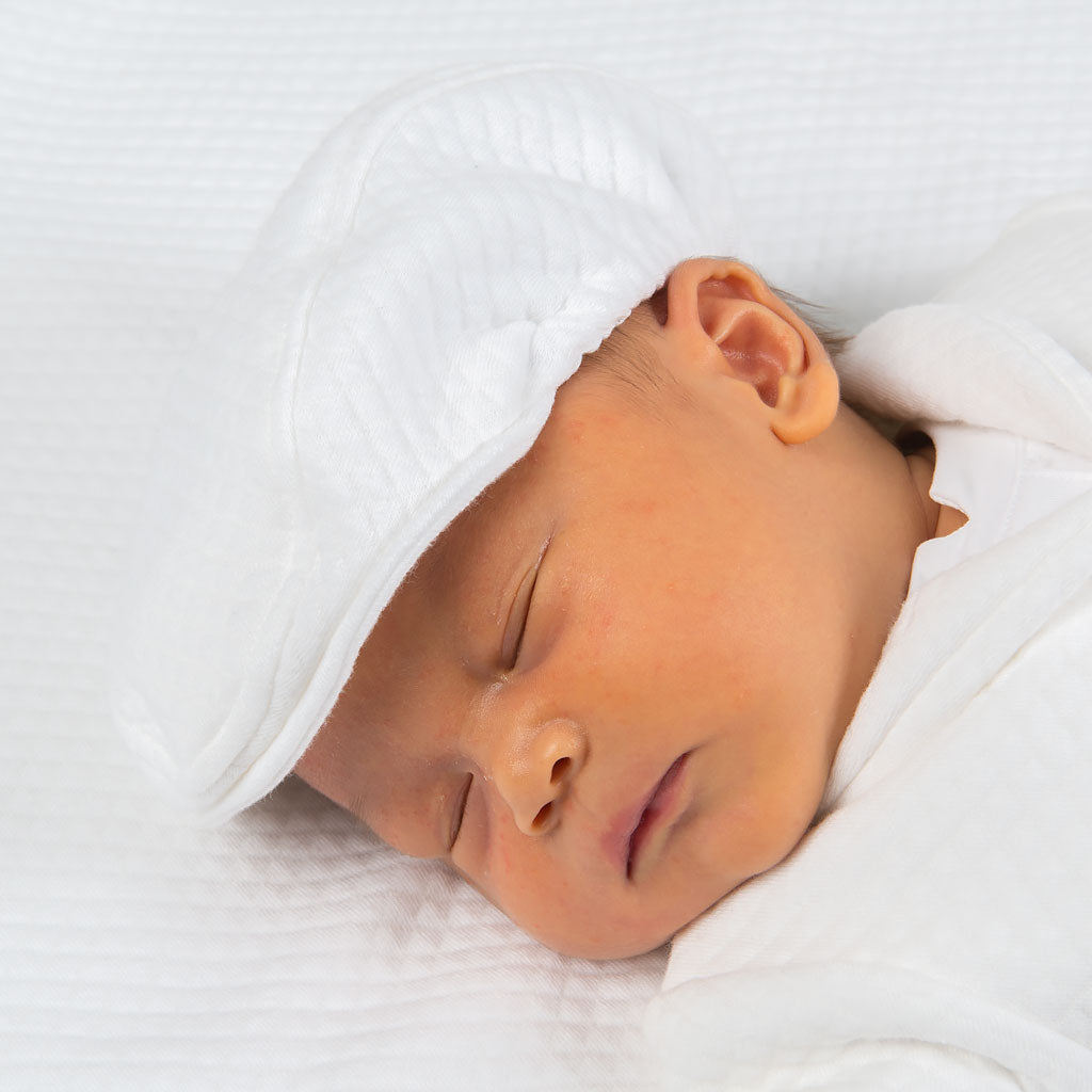 A sleeping baby, wrapped in a white blanket and wearing a cozy cap, rests peacefully on a soft surface in their charming Elijah Newborn 3-Piece Suit.