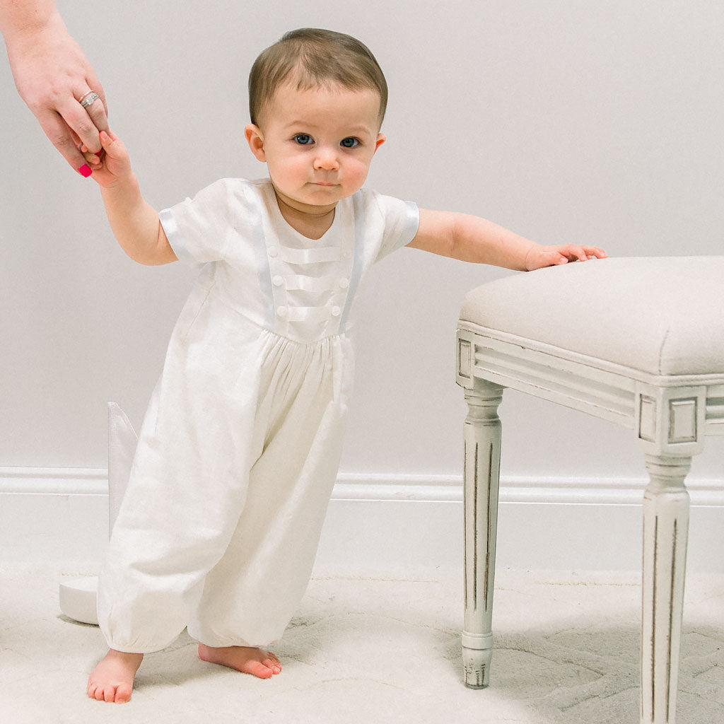 Baby boy standing up with the help of his mother's hand and wearing the Owen Linen Romper featuring silk ribbon in ivory and light blue across the front bodice and sleeves.