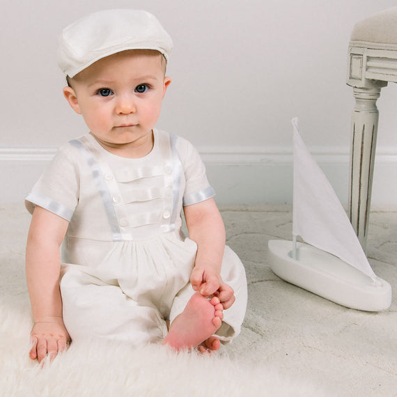 Baby boy sitting down and wearing the Owen Linen Romper featuring silk ribbon in ivory and light blue across the front bodice and sleeves