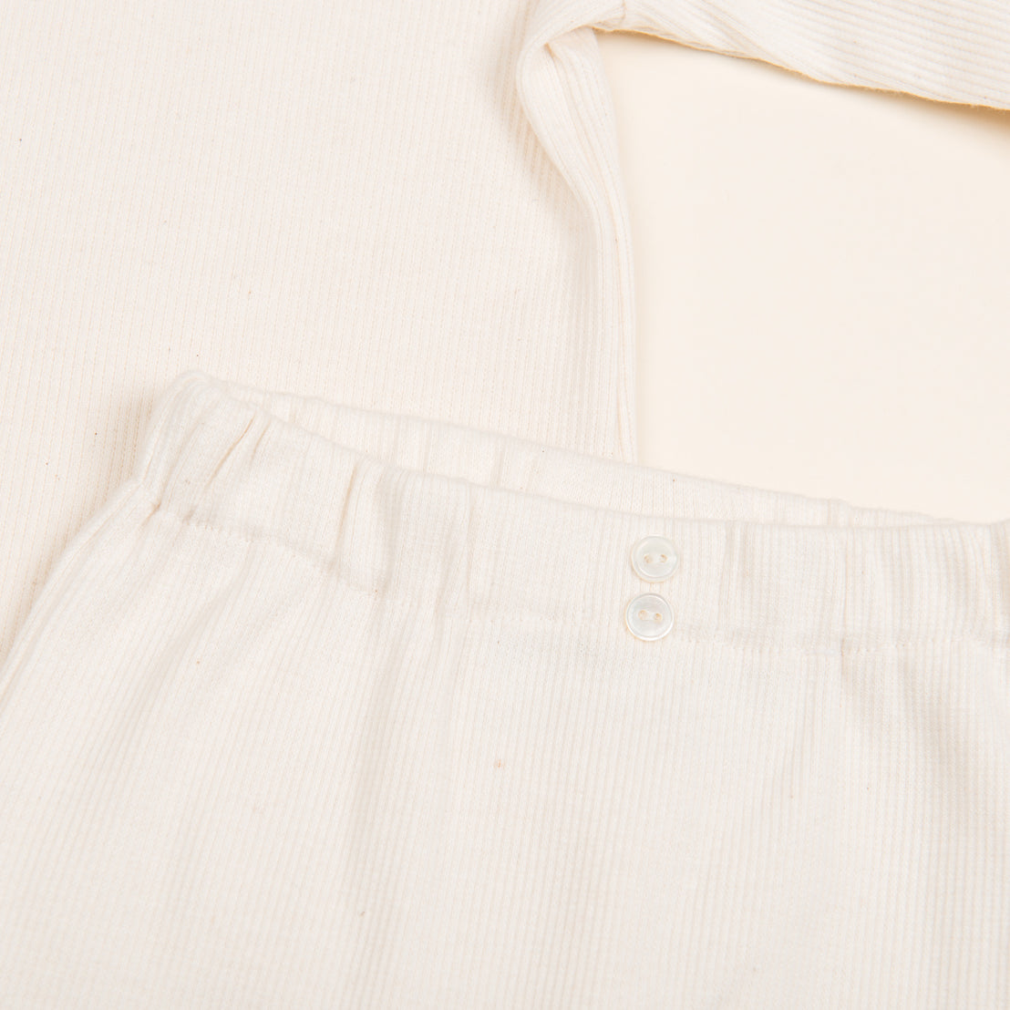 A close-up of the Natural Ivory Ribbed Cotton Top & Leggings, showcasing the cream-colored ribbed texture. The pants feature an elastic waistband with two small white buttons, paired with a matching long-sleeve top. Handmade in the USA, this outfit material looks wonderfully soft and comfortable.
