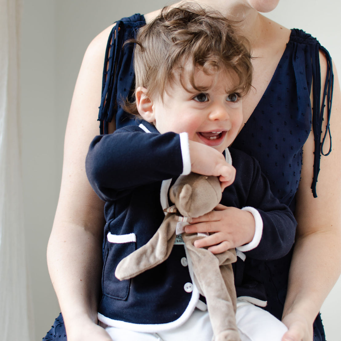 A smiling toddler with curly hair, wearing the handmade Elliott Jacket featuring a dark design with white trim, holds a stuffed bunny while sitting on an adult's lap. The adult wears a sleeveless navy French terry cotton dress, their heads not fully visible.