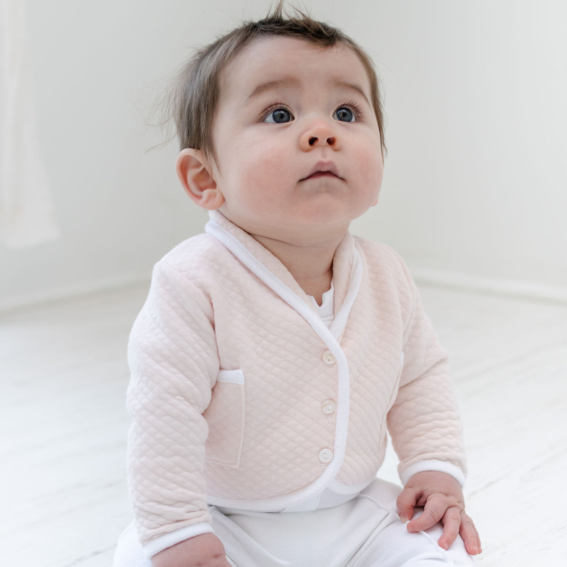 A baby dressed in the Asher Blush 3-Piece Shorts Suit sits on a white wooden floor, gazing upwards with wide eyes. The softly lit room creates a tranquil atmosphere.