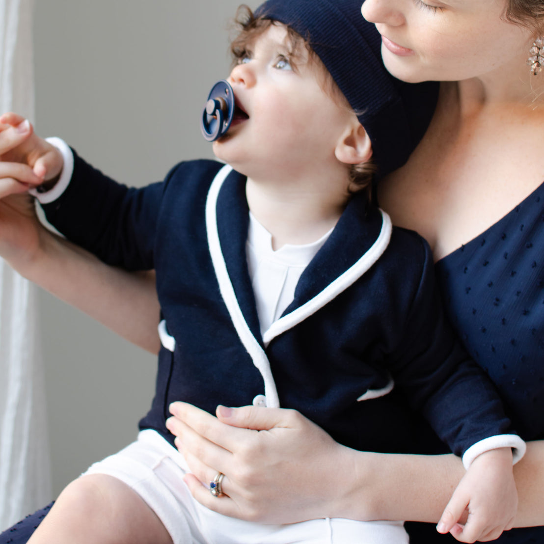 A woman holds a young child dressed in the Elliott Jacket, a navy French terry cotton sailor outfit, along with a pacifier. The child looks upward while sitting on her lap. The woman wears a navy dress adorned with a subtle dotted pattern and gently holds the child's hand.