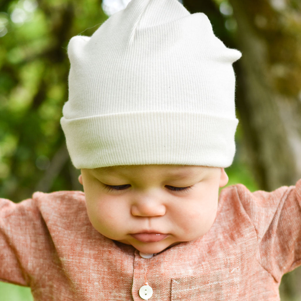 A baby sporting the Natural Ivory Ribbed Pima Beanie and a rust-colored shirt is looking down with their arms spread outward. The background features a blurred outdoor scene with green leaves and tree trunks, creating an enchanting setting for this handmade beanie.