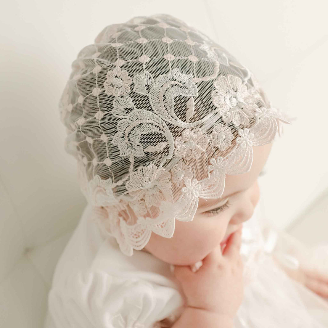 A close-up shot of an infant wearing a Joli Sheer Bonnet, gazing downward contemplatively with a finger near their mouth.