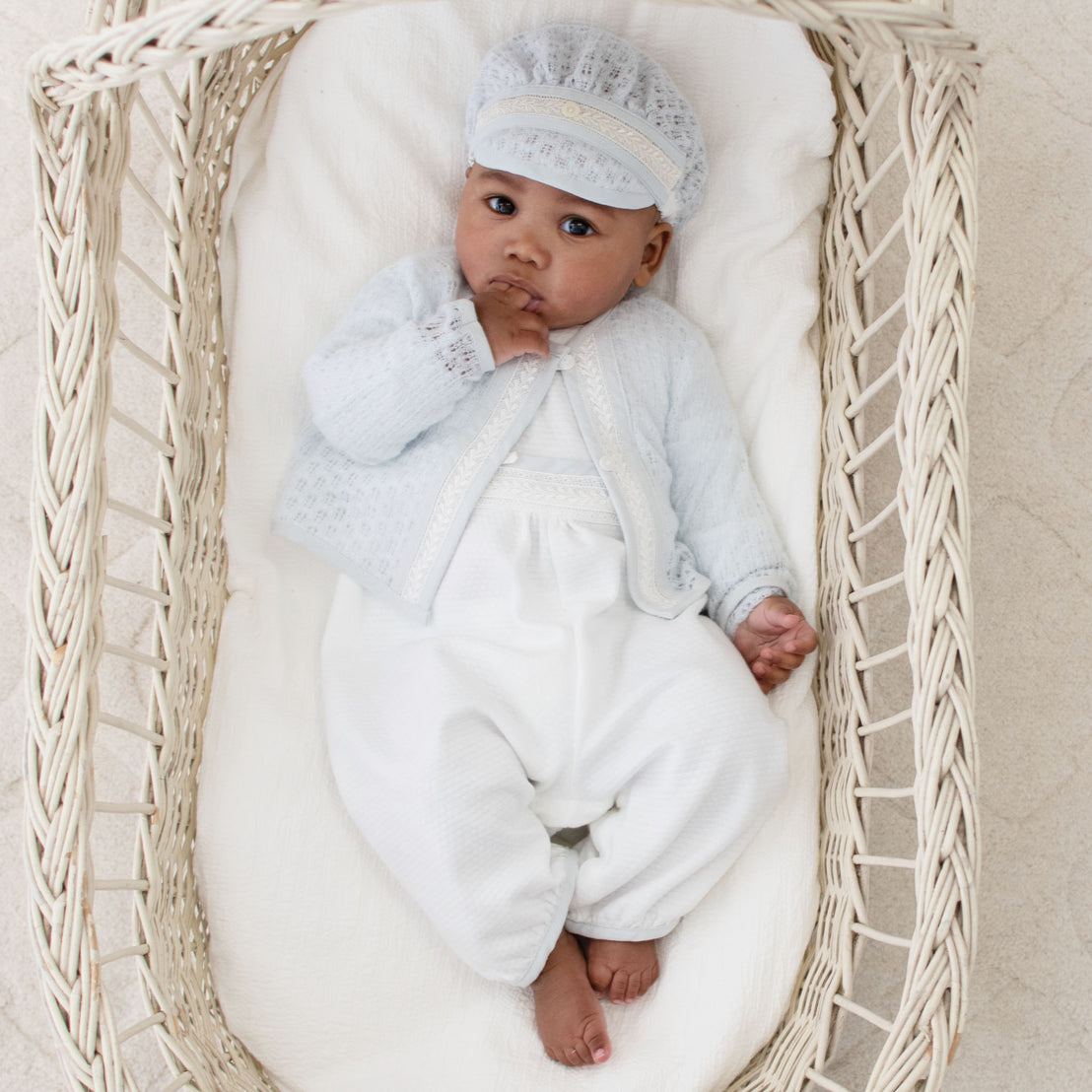 A baby laying in a crib and wearing the Harrison Long Sleeve Romper made from textured white cotton and featuring ivory Venice lace and a blue cotton/linen edging. The Harrison Long Sleeve Romper is paired with the Harrison Blue Knit Sweater and Hat
