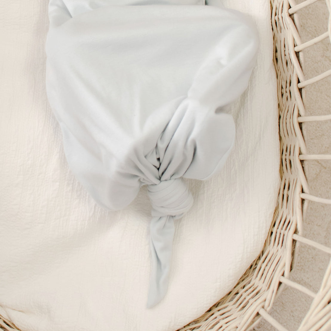 The Evan Knot Gown, made from 100% pima cotton in a light gray hue and handcrafted in the USA, is elegantly tied in a knot for a smooth, gathered look. Nestled inside a wicker basket on a textured white surface, this newborn boys' gown radiates softness and tranquility.
