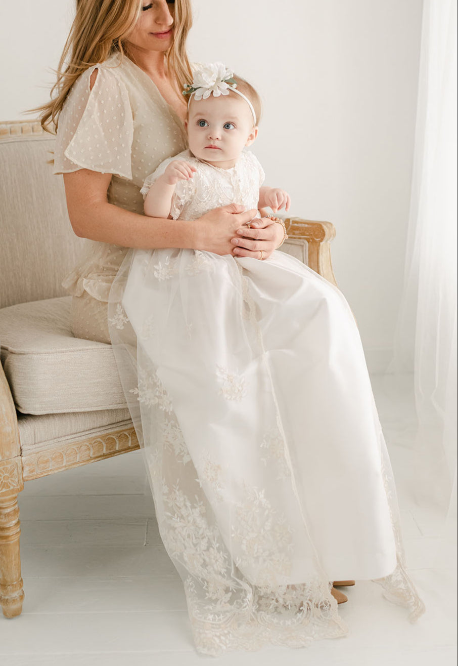 A woman sits on an elegant armchair holding a baby dressed in a white lace gown and headband for a christening, both looking towards the camera in a bright room