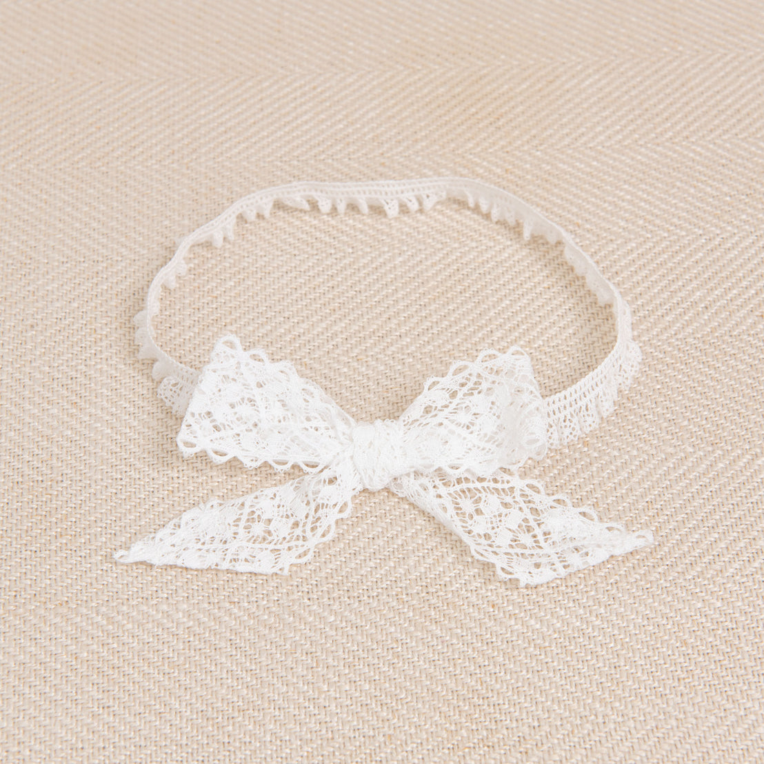 A Thea Lace Headband, a delicate lace bow with light ivory stretch lace, displayed on a textured beige fabric background.