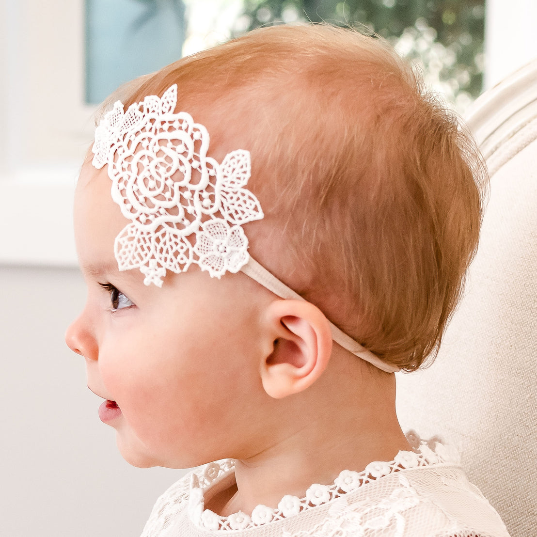 Profile view of a baby with light hair wearing a delicate Juliette Lace Headband with a large floral design, sitting indoors against a soft, bright background.