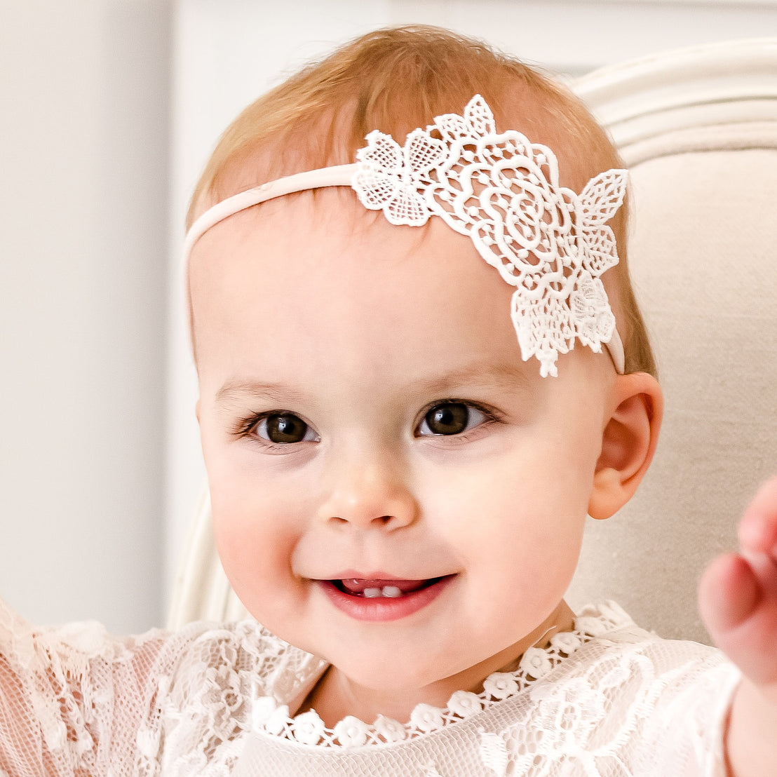 A smiling baby wearing a Juliette Lace Headband, sitting in a chair and reaching out toward the camera.