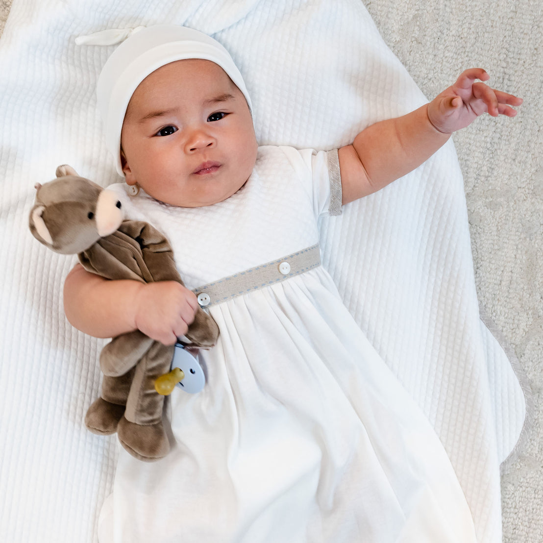 A baby dressed in a boutique white dress and headband lies on a soft blanket, holding an Austin Bear Buddy pacifier holder, looking at the camera with a gentle expression.