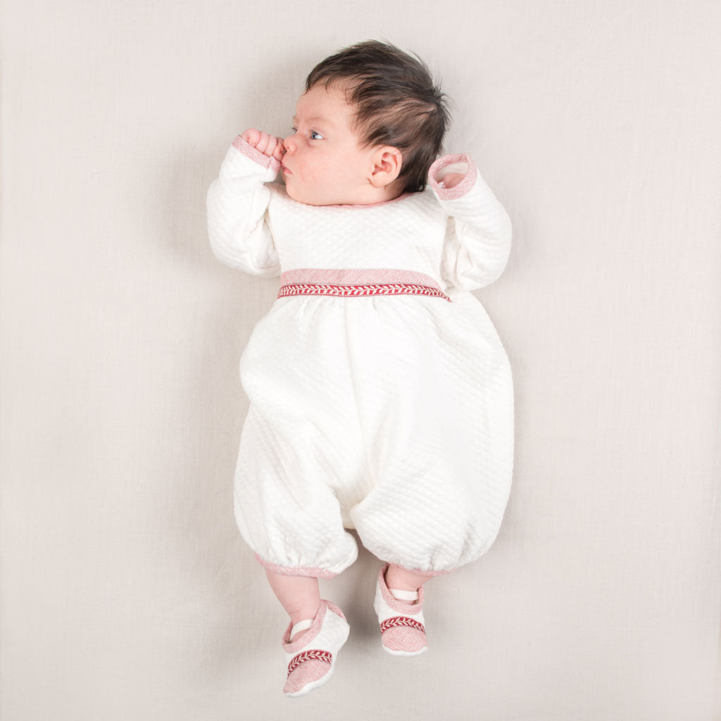 A baby wearing the Sweetheart Newborn Romper, made from 100% quilted cotton with red trim, lies on a light surface. The baby gazes to the side with one hand near their mouth while sporting matching red and white shoes.