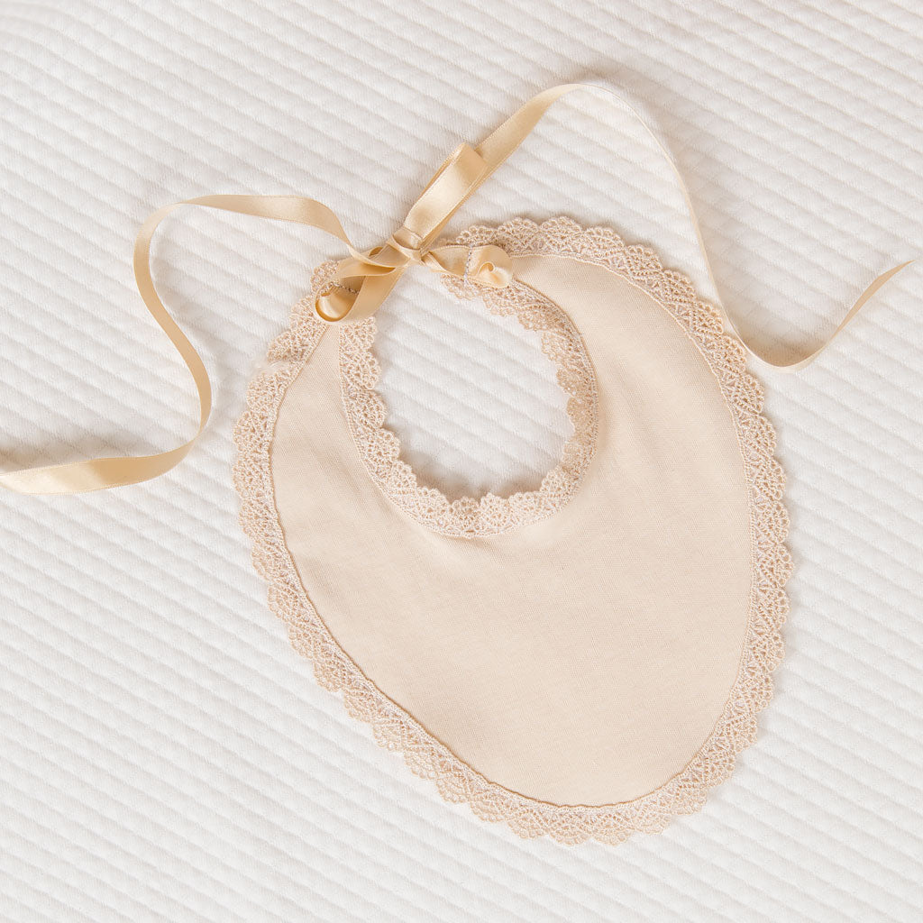 The Mia Newborn Bib made from a champagne pima cotton with intricate matching lace detailing around the edges and gold silk ribbon ties. The bib is photographed on a white quilt fabric background.