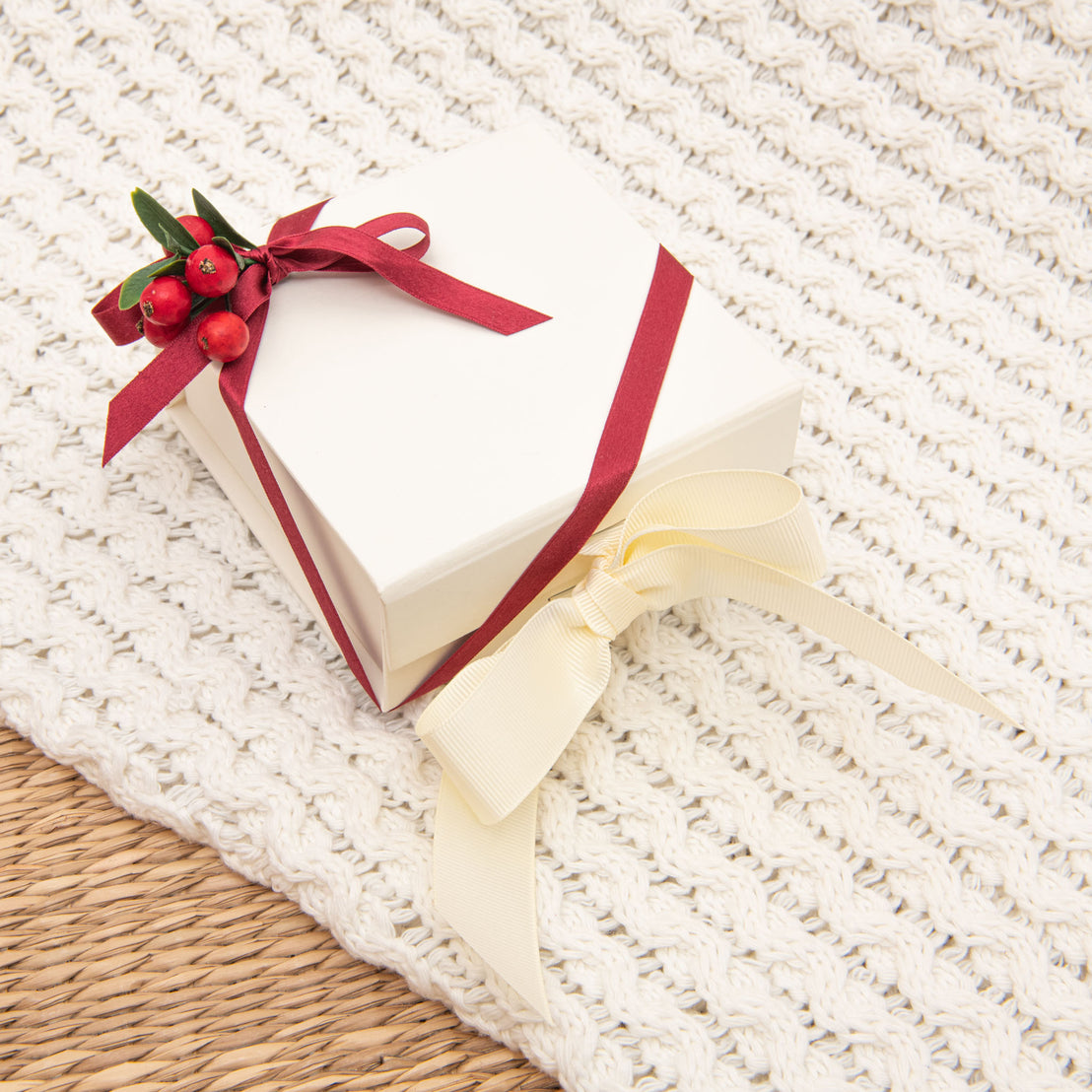 A square white Baby Beau & Belle Christmas Gift Wrap adorned with a red ribbon and a sprig of holly on a cream knitted background, positioned partially on a woven mat. This box features magnetic closure, enhancing its elegance.