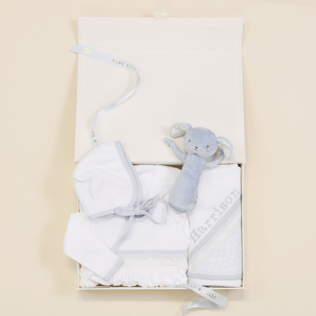 The Harrison Newborn Gift Set box showing the gown, plush bunny rattle, personalized blanket, and hat, elegantly presented with a Baby Beau & Belle ribbon.