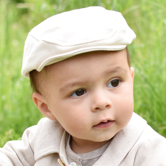 A baby wearing a Natural French Terry Newsboy Cap and a soft French Terry Cotton outfit is sitting outdoors on the grass. The curious little one gazes to the side, captivated by the world around them.