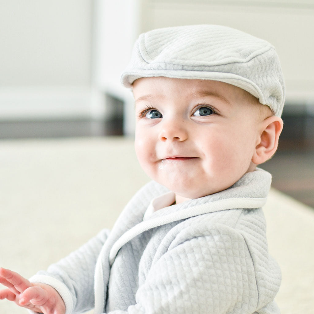 Baby boy smiling into camera wearing the quilted cotton newsboy cap, part of the Asher formal baby outfit collection.