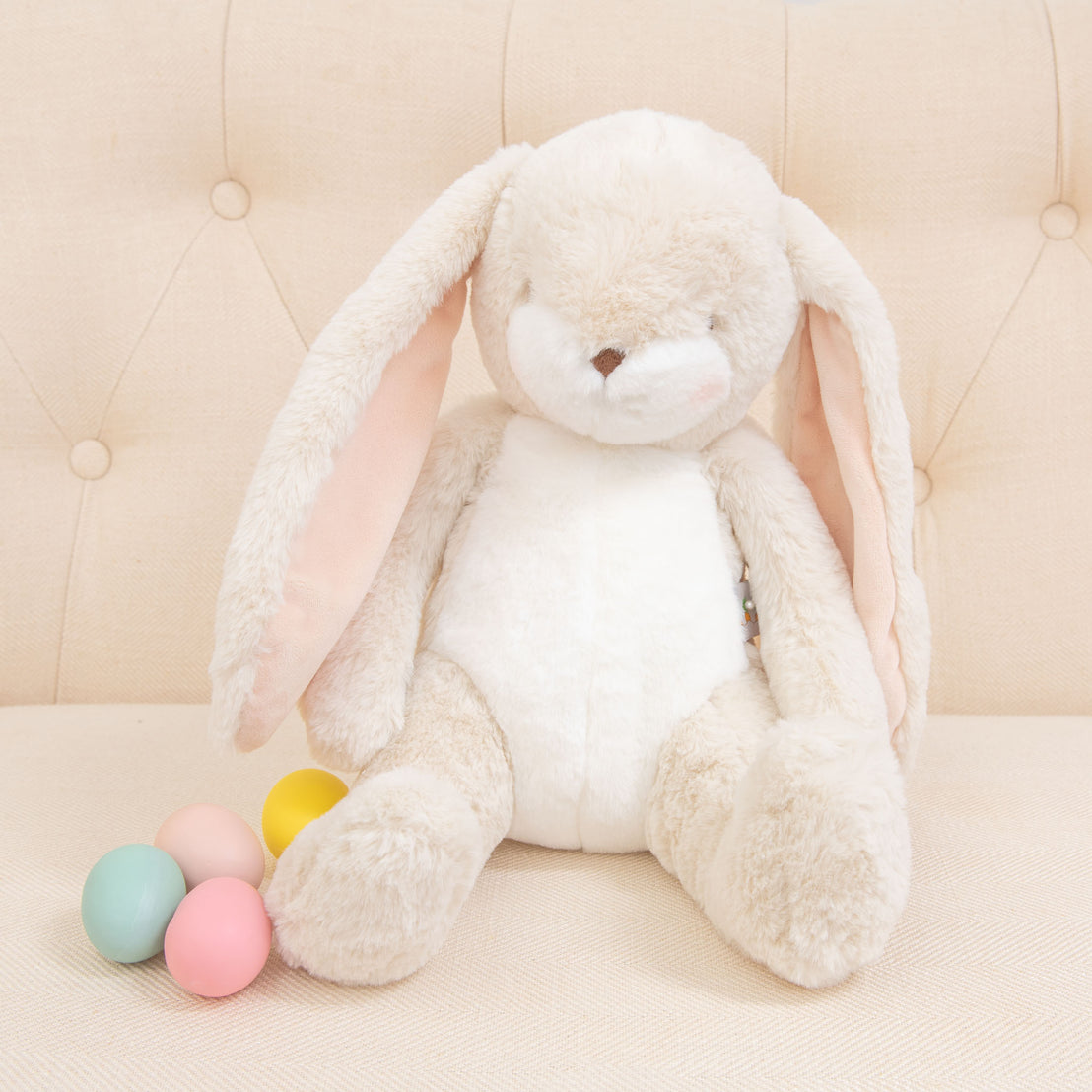 A Cream Nibble Bunny, specifically designed to be baby-safe and soft, sits on a beige tufted couch with its long ears. Next to it sit four pastel-colored eggs in pink, yellow, blue, and green, completing this adorable scene.