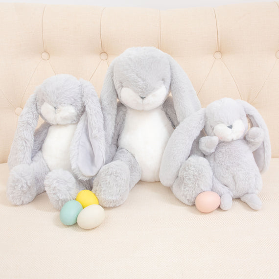 On a beige sofa, three Grey Nibble Bunnies are seated, each adorned with a colorful egg—one yellow, another green, and the last pink—placed before them. These baby-safe soft plush bunnies boast floppy ears and an embroidered face, radiating gentle charm through their soft and fluffy textures.