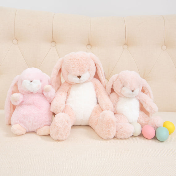 Three Pink Nibble Bunny toys in shades of pink and white sit on a beige, tufted sofa. They are surrounded by pastel-colored eggs, creating a soft and cozy scene.