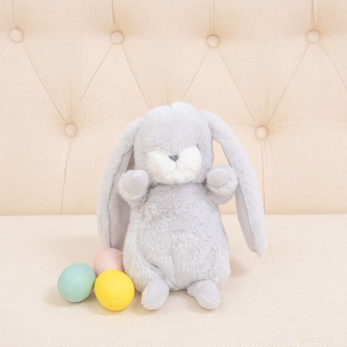 A fluffy Grey Nibble Bunny with an embroidered face and long ears sits on a beige tufted cushion. Next to it are four pastel-colored eggs: pink, green, blue, and yellow. The soft and neutral background enhances the cozy atmosphere of these baby-safe soft plush companions.