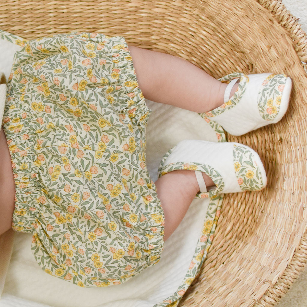 A baby wearing Petite Fleur Bloomers with matching shoes lies in a handcrafted wicker basket, with their legs playfully bent and visible from the top.