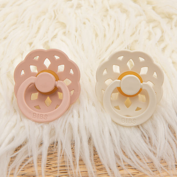 The Ava Pacifier Set that includes two pacifiers, one in blush and the other in ivory, with round, cut-out shields, displayed on a on a white fluffy textured surface.