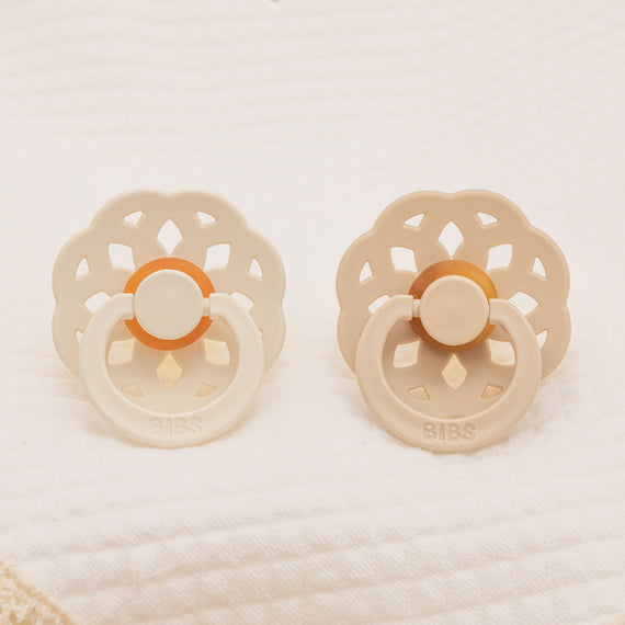 The Chloe Pacifier Set in Ivory & Vanilla consists of two floral-shaped pacifiers resting on soft, textured white fabric. Each pacifier is crafted from natural rubber and manufactured in Denmark, featuring an intricate cut-out design on the handle in neutral shades of ivory and vanilla. The brand name "BIBS" is visible, making them perfect for helping babies self-soothe.