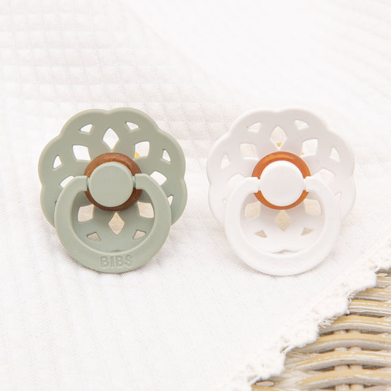 Two Eliza Pacifier Sets, one sage green and one white, on a soft white cloth with a woven pattern at the corner, perfect for christening.